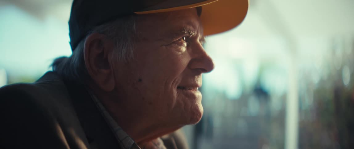 A close view from the side shows an elderly veteran wearing a black and yellow cap while smiling and looking out a window as soft light illuminates his face