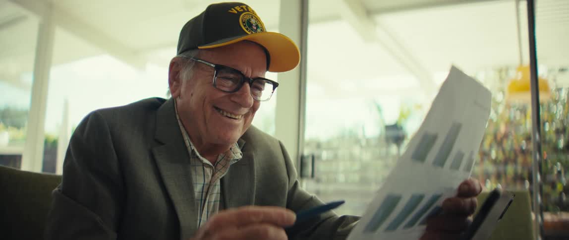 An elderly man wearing a veterans cap smiles warmly while reviewing a document with bar chartshe eventually takes off his glasses and laughs looking pleased with the information he is reading