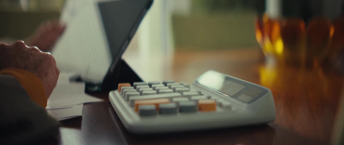 Elderly person uses a calculator at home while referencing papers and a computer