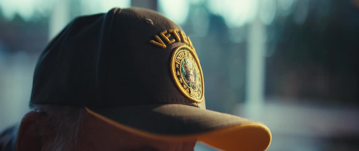 A close up profile shot of an elderly man wearing a dark green veteran cap featuring a detailed gold united states army emblem embroidered on the front while soft natural light filters through a blurred background