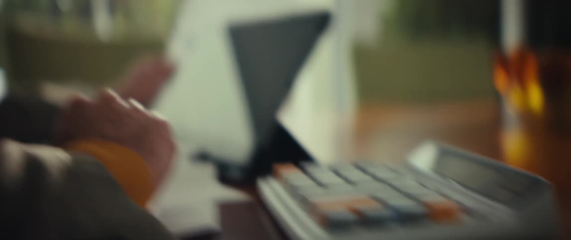 A close view shows a man wearing glasses and a veteran hat as he looks at papers and a tablet with a white calculator on a desk in a bright room