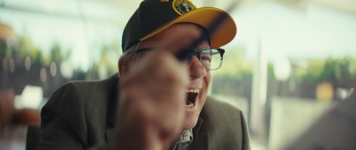 Elderly man wearing glasses and a cap laughs while using an electronic device indoors