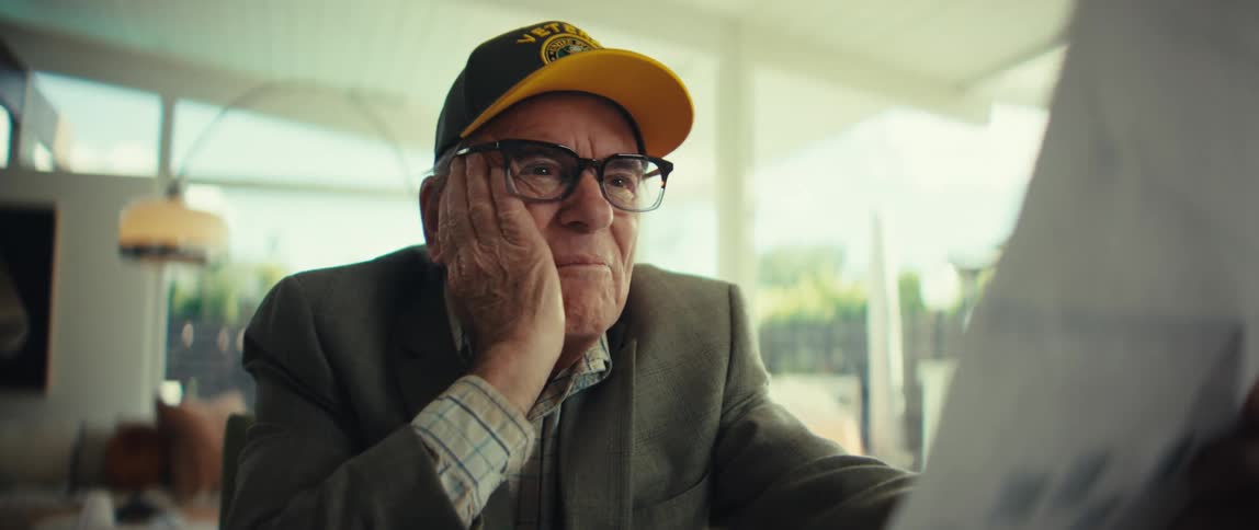 Elderly man wearing a cap and glasses raising his hand while reacting as he looks at a sheet of paper in a welllit indoor setting