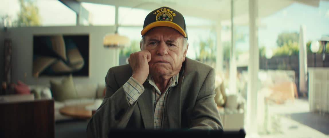 An elderly veteran wearing a black army cap and a grey suit jacket removes his glasses and rests his chin on his hand while looking at a tablet in a bright living room