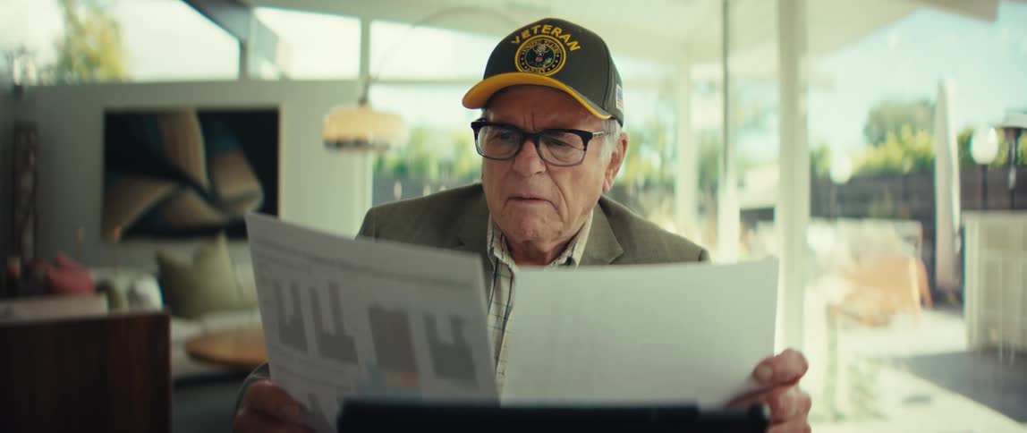 Elderly man wearing a cap and glasses holding two sheets with charts while reviewing documents in a room with large windows in the background