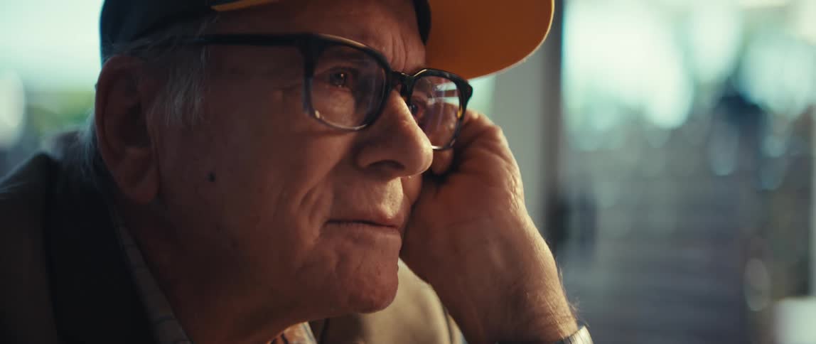 The video shows a closeup profile of an elderly man wearing glasses and a yellow baseball cap he rests his head on his hand with a pensive expression as he gazes offcamera