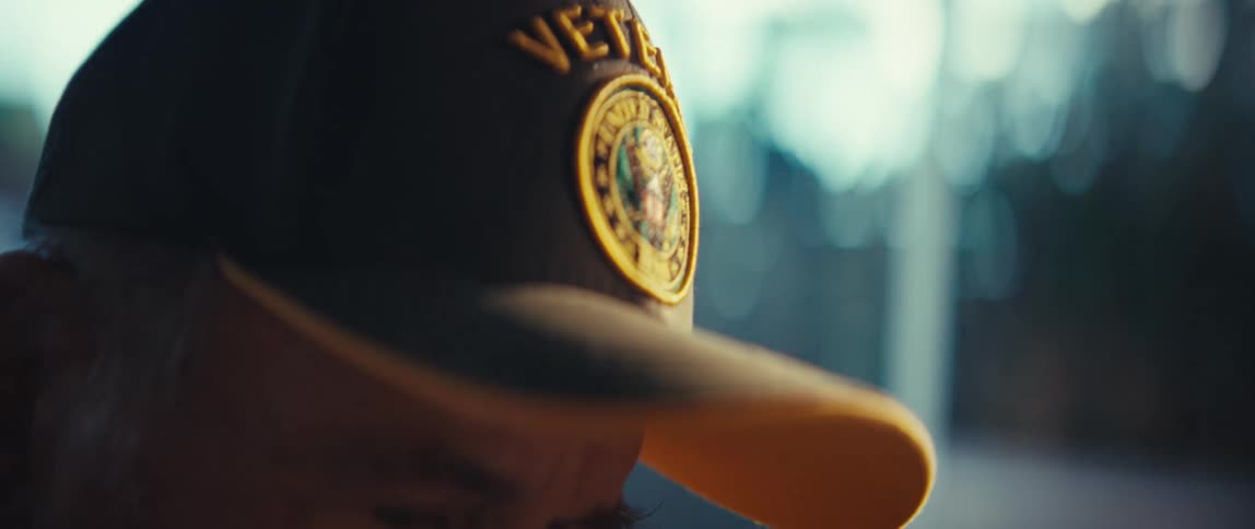 A close view shows the gold embroidered veteran patch on a black and yellow cap worn by an elderly man who looks down with a serious expression in a brightly lit indoor space
