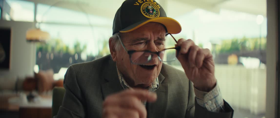 Elderly man wearing a cap and glasses looking at a sheet of paper with an attentive expression in a welllit indoor setting