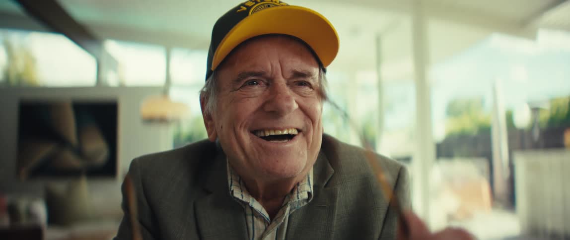 Elderly man wearing glasses and a cap smiles while using an electronic device indoors