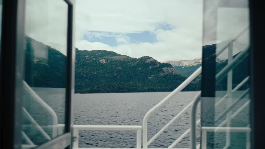A view through the open glass doors of a boat shows a wide lake with dark water and green mountains under a cloudy sky