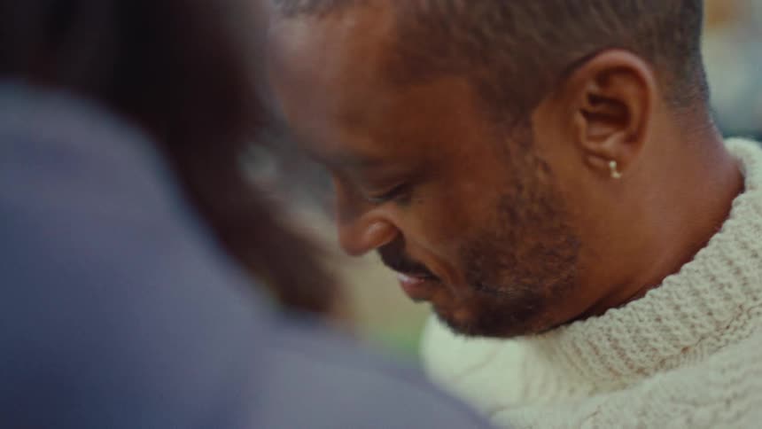 A smiling happy black man is seen from the chestsitting next to a partially obscured person outdoors