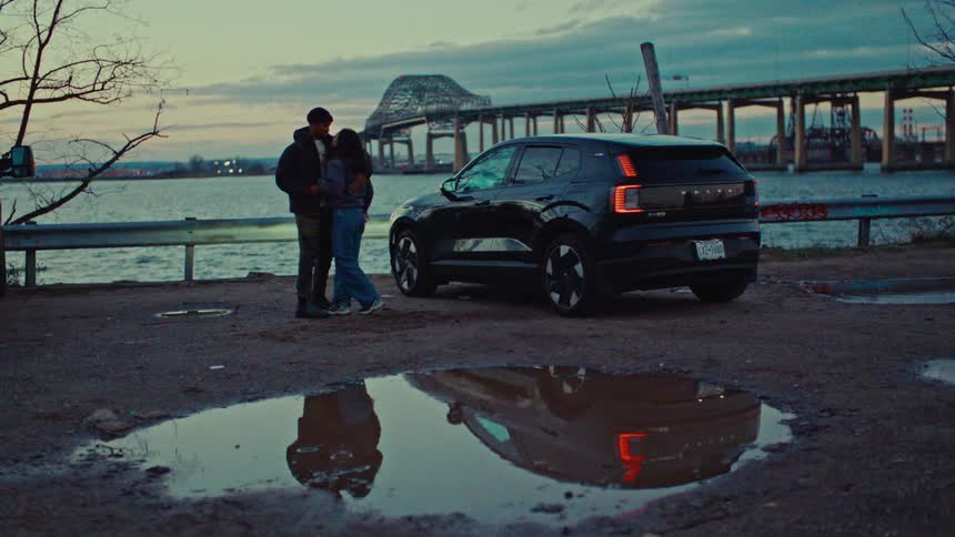 Couple embracing beside a parked car by the river with a bridge in the background and reflection in a puddle