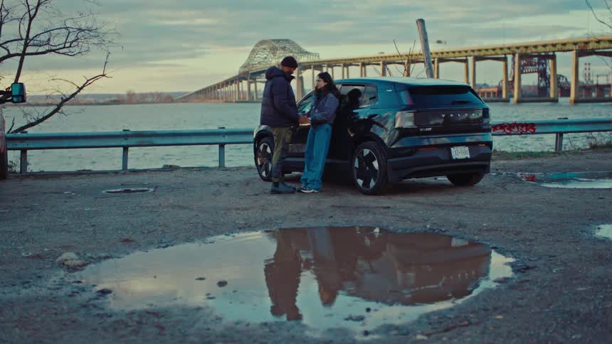 A man an woman talk while standing next to a car parked by a river