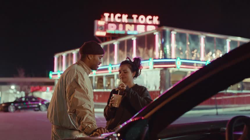 Couple sharing a dessert beside a car in front of a diner with a neon sign at night