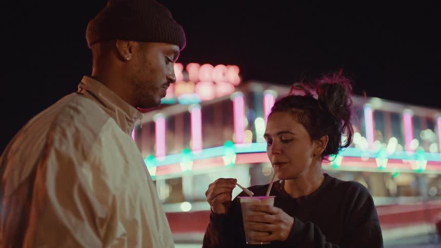 A man and woman stand by a car at night in front of a neon diner and the woman is holding a milkshake playfully feeding a fry to the man while they smile and talk together in the colorful ambient light