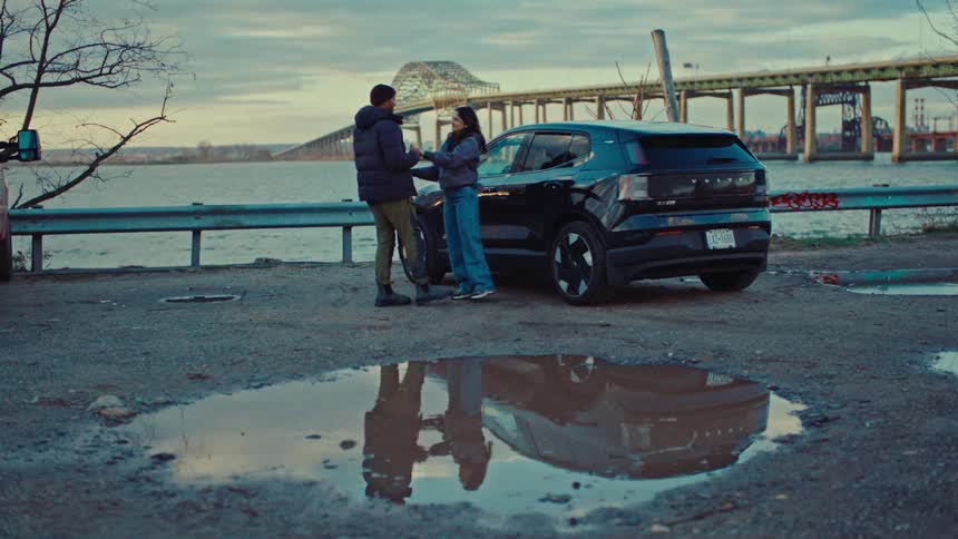 Couple dances near a car parked in a waterfront parking lot containing reflective pools