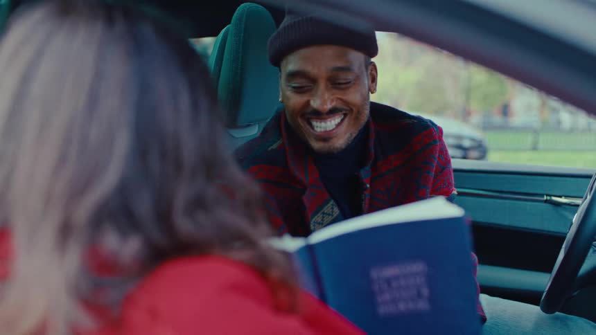 A man with a beanie sits in the driver seat laughing while looking at a blue book held by a woman in a red jacket