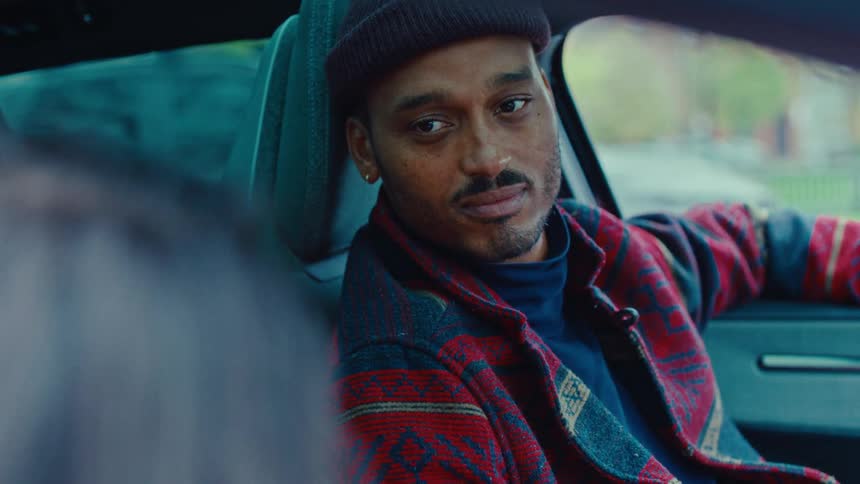 A man wearing a beanie and patterned red jacket sits in the drivers seat of a car looking thoughtfully toward someone beside him