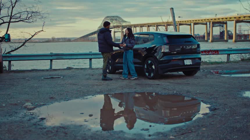 Couple embracing beside a parked car near a bridge over the river with reflection in a puddle