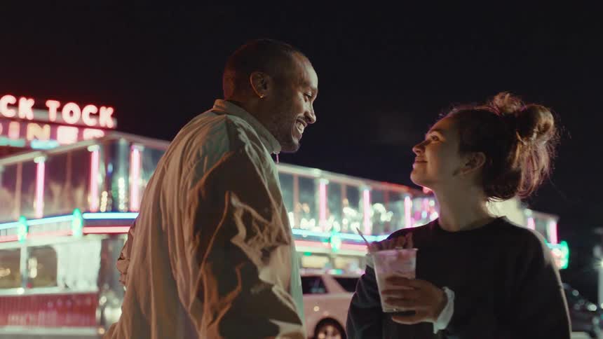 A couple shares a playful moment at night in front of a brightly lit dinerthe woman holds a milkshake and playfully dabs some on the mans nose before they lean in close and laugh together