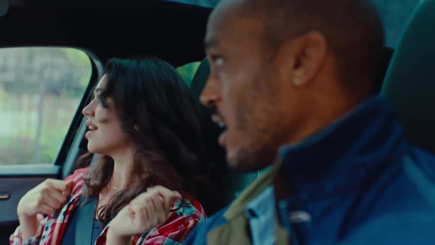 A woman wearing a red plaid shirt sings and dances in the passenger seat of a car while a man in a blue jacket drives during a road trip
