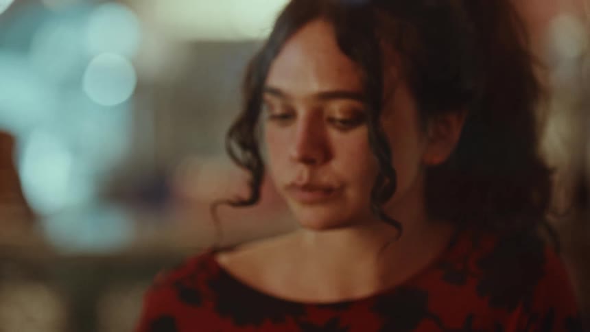 A woman with curly brown hair wearing a red floral shirt eats with a spoon while sitting in a dimly lit restaurant with blurred lights in the background