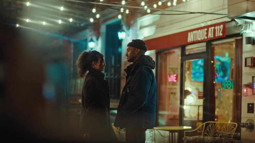 A dimly lit urban street scene at night with a focus on a couple standing smiling and talking