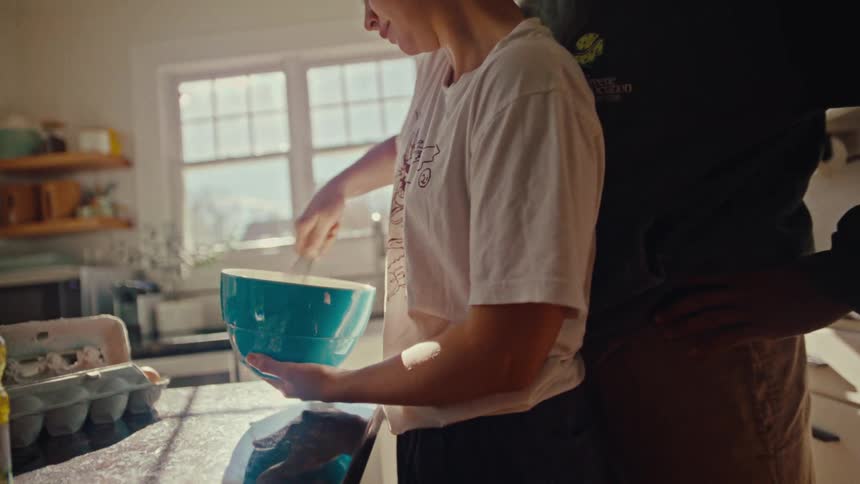 A woman vigorously whisks ingredients in a bright blue bowl while a man stands closely behind her in a sundrenched kitchen watching the preparation as warm morning light pours through a large window onto the granite counter