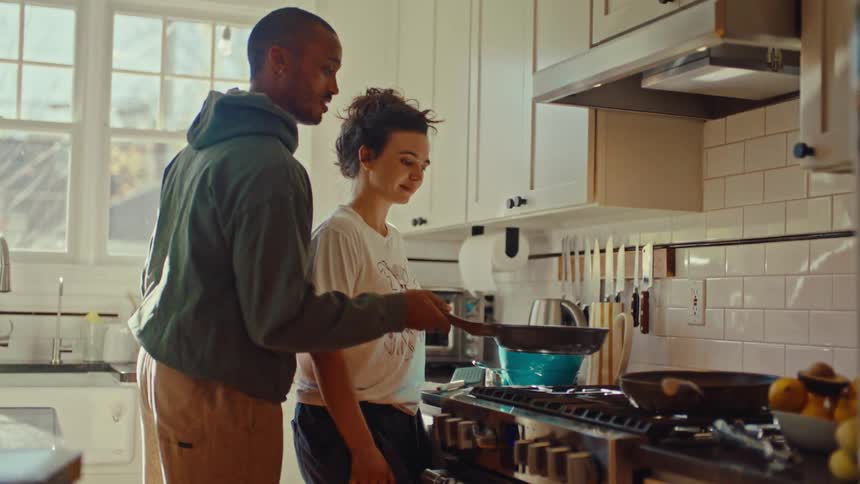 A young man and woman stand together in a brightly lit kitchen smiling while the man helps the woman toss food in a frying panthe couple appears relaxed and happy as they share the task of cooking over a professionalstyle gas stove