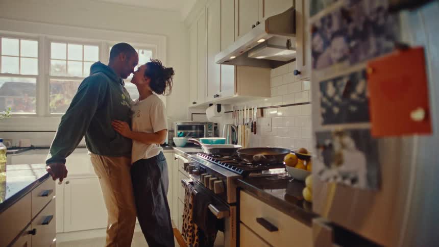 A man wearing a green hoodie and a woman wearing a white t shirt kiss and laugh while hugging and kissing in a bright kitchen during the day