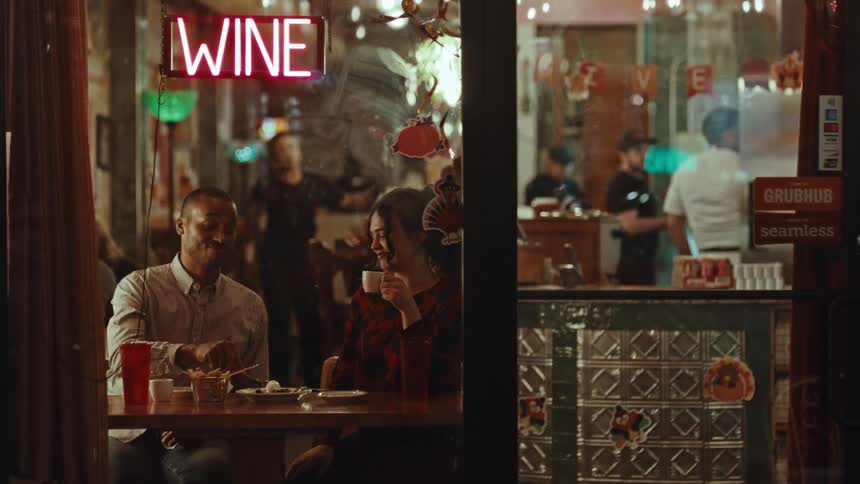 Young adult woman sitting and talking while eating dessert and having coffeeat a restaurant with other customers and servers on the background seen through a window during night time