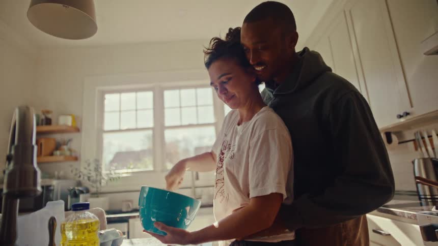 Man hugging a woman while she mixes ingredients in a bowl in the kitchen