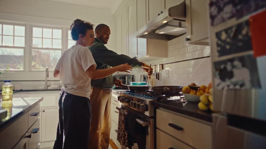 A man wearing a green hoodie and a woman wearing a white t shirt prepare food together at a stove in a bright kitchen during the day