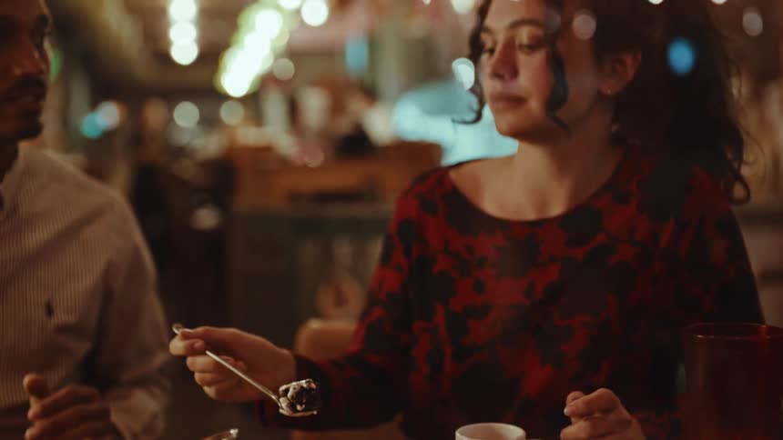 A woman in a red floral dress eats a large brownie topped with vanilla ice cream while sitting at a restaurant tableher companion watches her take a bite and she smiles as she enjoys the dessert during their conversation