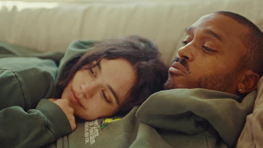 A man and woman wearing hoodies relax together on a couch
