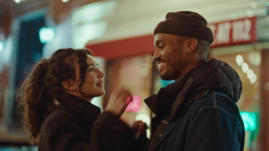 A man wearing a beanie and a woman in a black coat look at each other and kiss while standing on a city street at night with colorful lights in the background