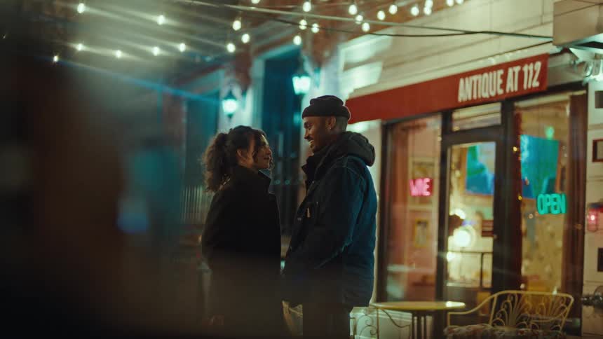 Under a canopy of warm cafe lights a couple shares an intimate moment on a chilly city sidewalk they lean in close laughing and touching faces before finally embracing for a romantic kiss