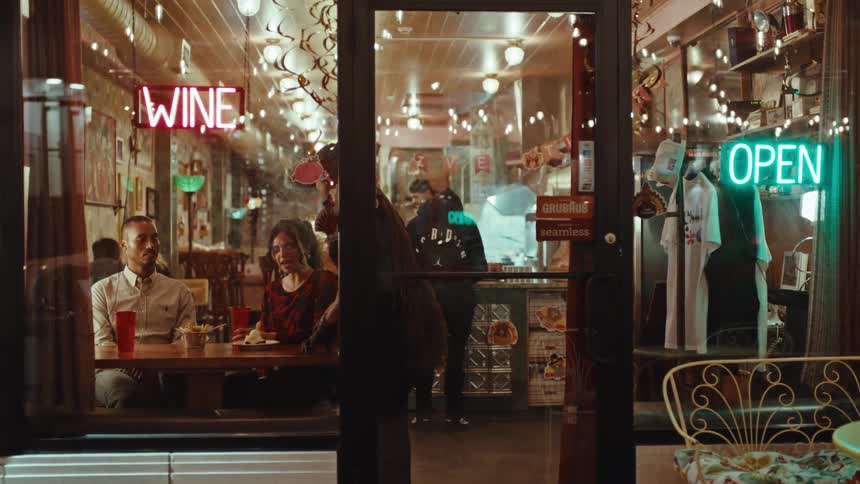 A man and a woman on a date sit at a table inside a brightly lit restaurant at night while a worker serves them food through a glass door