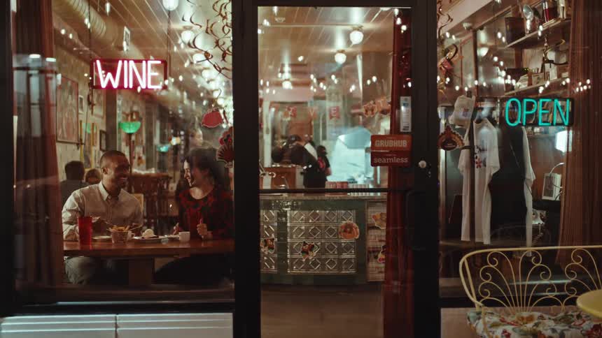 A man and a woman on a date sit at a table inside a brightly lit restaurant at night while a worker serves them food through a glass door