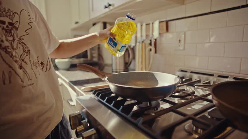 A person stands in a kitchen and carefully pours cooking oil from a large plastic bottle into a frying pan on the stovethe scene focuses on the golden liquid flowing into the pan while another person stands in the background watching