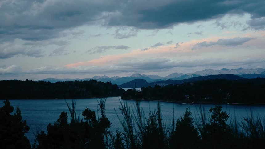 A view of a calm lake surrounded by dark hills and distant mountains under a cloudy sky during twilight