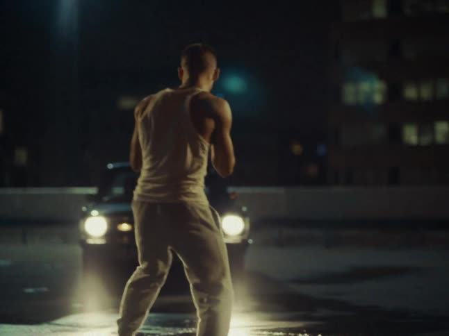 Young adult man wearing an undershirt shadowboxing in a parking lot in front of the headlights of a car in a metropolitan area outdoors during night time