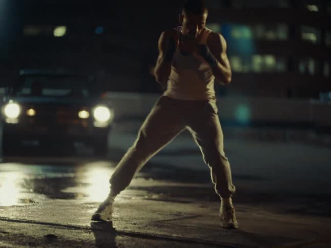 A man in athletic wear and hand wraps practices shadowboxing on a dark urban streetthe headlights of a parked car illuminate his movements against the backdrop of city buildings at night