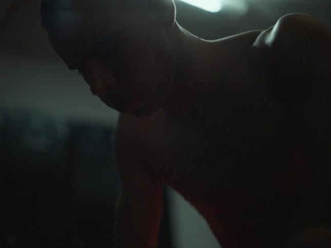 A shirtless man with short hair strains and grimaces while performing physical exercise in a dark gym with a single overhead light casting shadows on his face and shoulders
