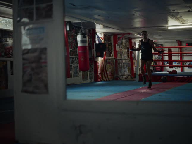 A man wearing a black tank top and shorts jumps rope in a boxing gym with a ring and a red punching bag in the background