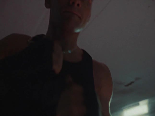 A male boxer wearing a black tank top and hand wraps practices shadow boxing inside a gym with a red floor and heavy bags throwing a quick series of punches toward the camera while moving his feet and upper body in a focused training session