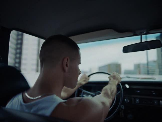 A man with a buzz cut wearing a white tank top drives a car through a city