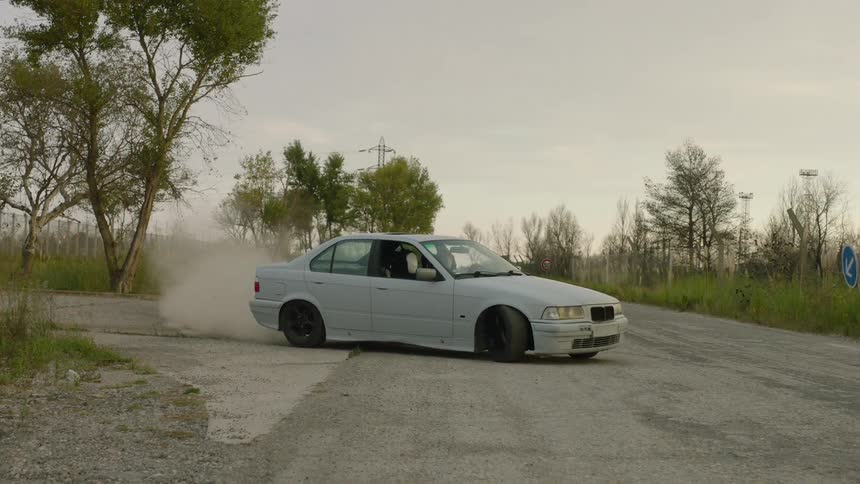 A light colored clear grey sedan performs a controlled drift onto a paved road kicking up a significant cloud of dust from the shoulder the car then continues driving forward past a fenced industrial area under an overcast sky