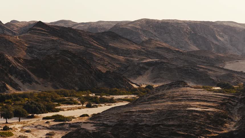 A wide view shows a vast desert landscape with jagged dark rock formations and a dry valley filled with scattered green trees and sand under a bright hazy sky