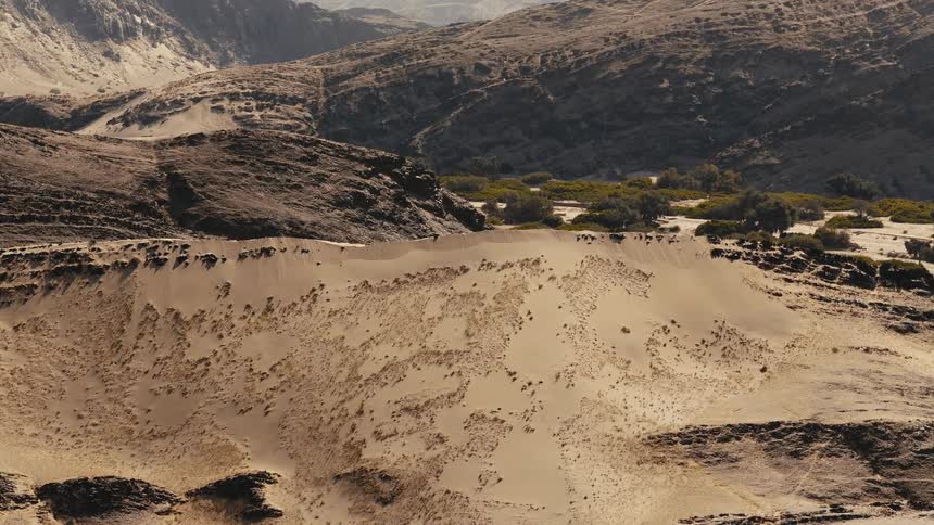 A massive sand dune nestled within a rugged mountain range where sparse green vegetation dots the base of the arid slopes under a bright sun that casts deep shadows across the vast and desolate desert floor
